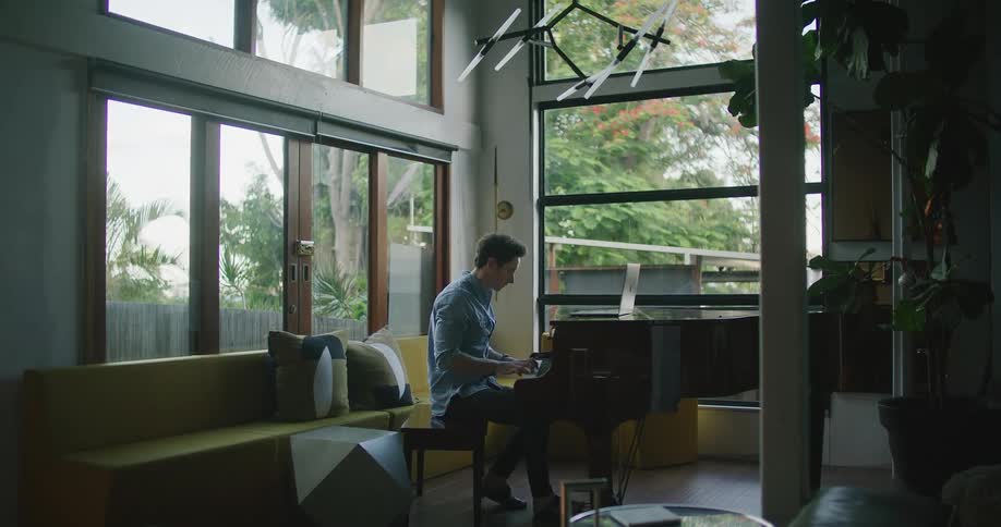 A man in a room sitting and playing a piano with a laptop on top of it