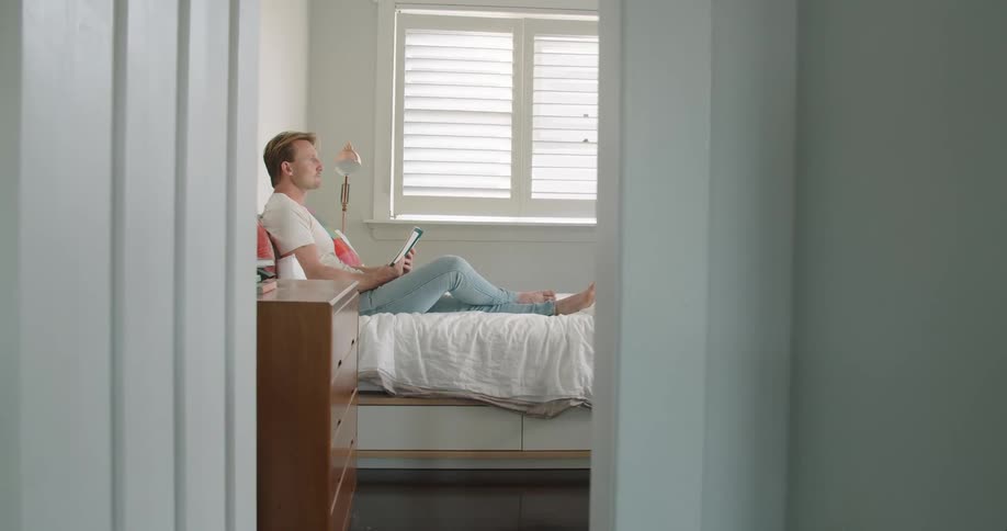 A doorway view of a man sitting on a bed in a room reading a book at daytime