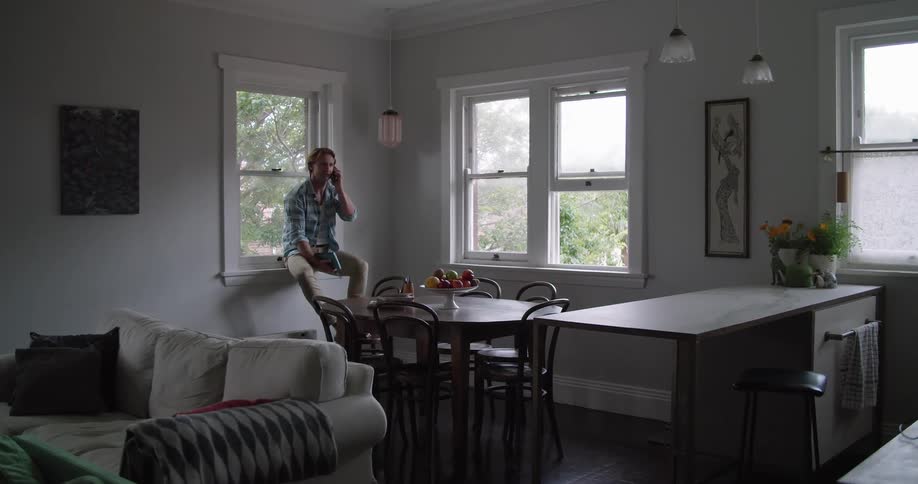 A man talks on the phone while sitting on a window sill with his feet up on a dinette chair