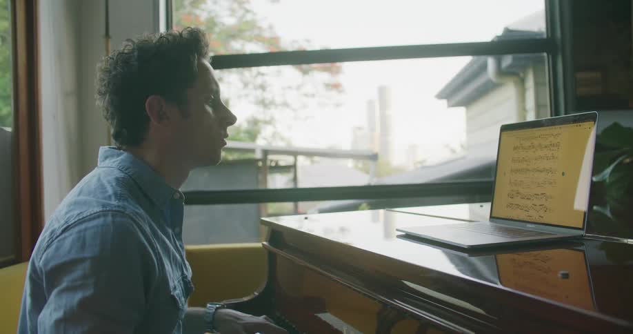 A male pianist reading sheet music on a laptop while playing a piano