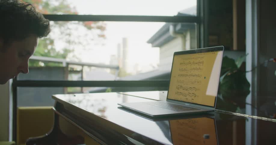 A pianist plays a piano while glancing at a music sheet being displayed on the screen of a laptop