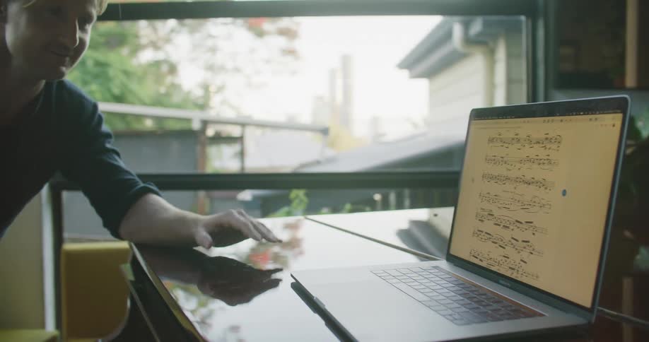 Two men in a room by a piano next to a laptop with a door and a window in the background