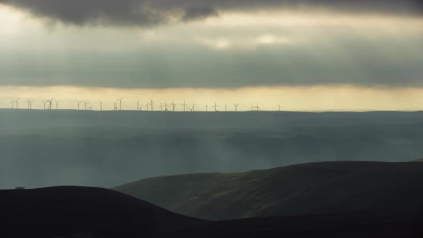 Dappled storm light floods into this wide aerial shot looking across a vast canyon towards hundreds of windmills at a large wind farm as they rotate creating clean renewable energy
