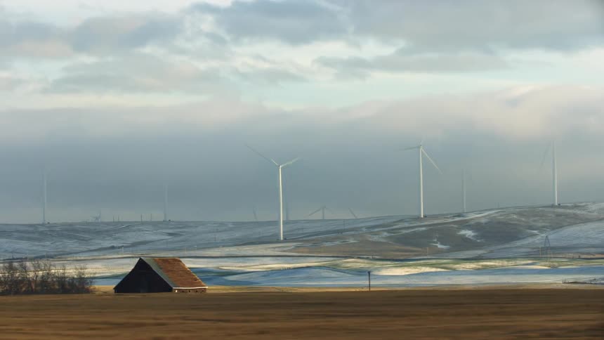 Low altitude aerial shot of rural farm land and snow covered hills with power generating wind mills rotating high on the ridge