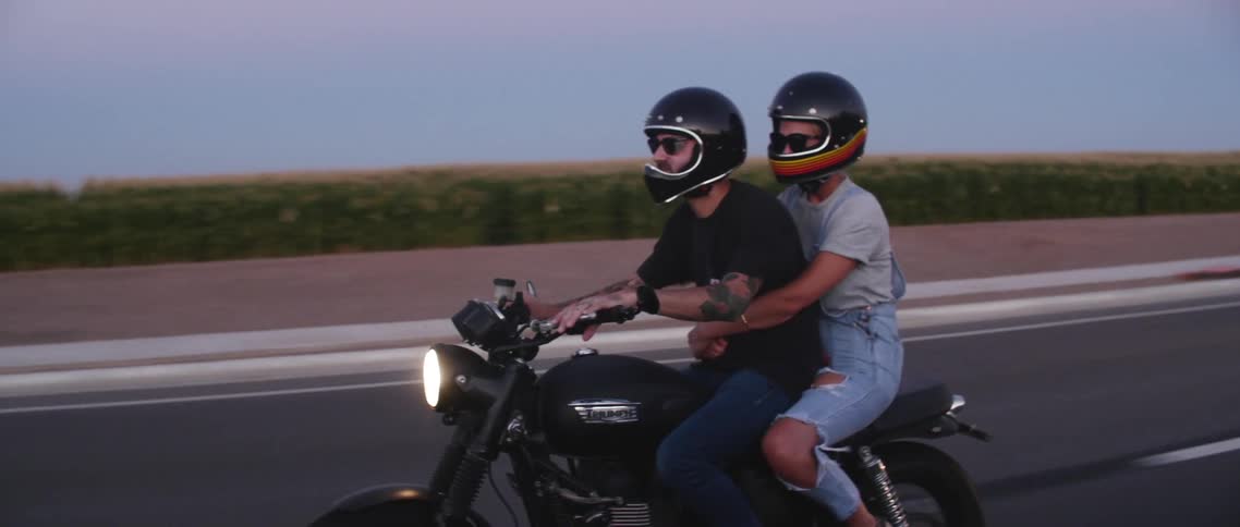 Two people with helmets riding a motorcycle with headlights on a road