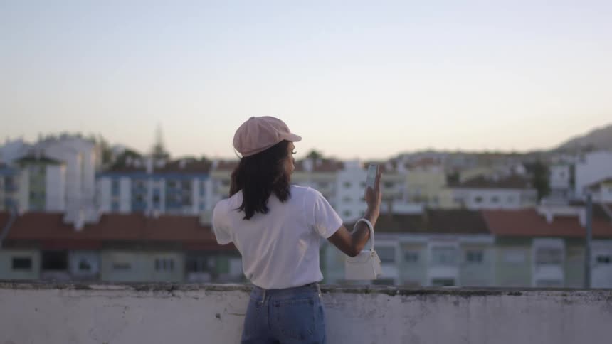 A woman standing outside across from a set of buildings takes a selfie with a smart phone
