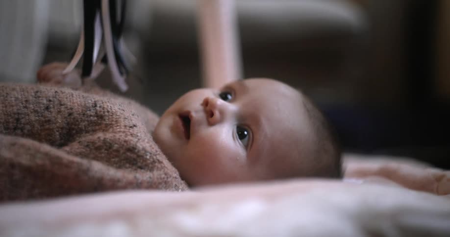 A young baby lays on its back while wiggling as it looks up at some dangling ribbons with great amusement