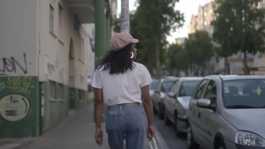 A woman walks down on a sidewalk in a city next to parked vehicles and looks around