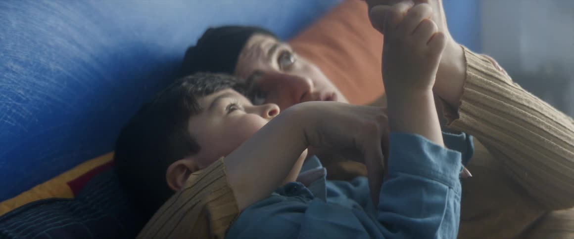 A woman lays on a bed with her small son and they play with their hands