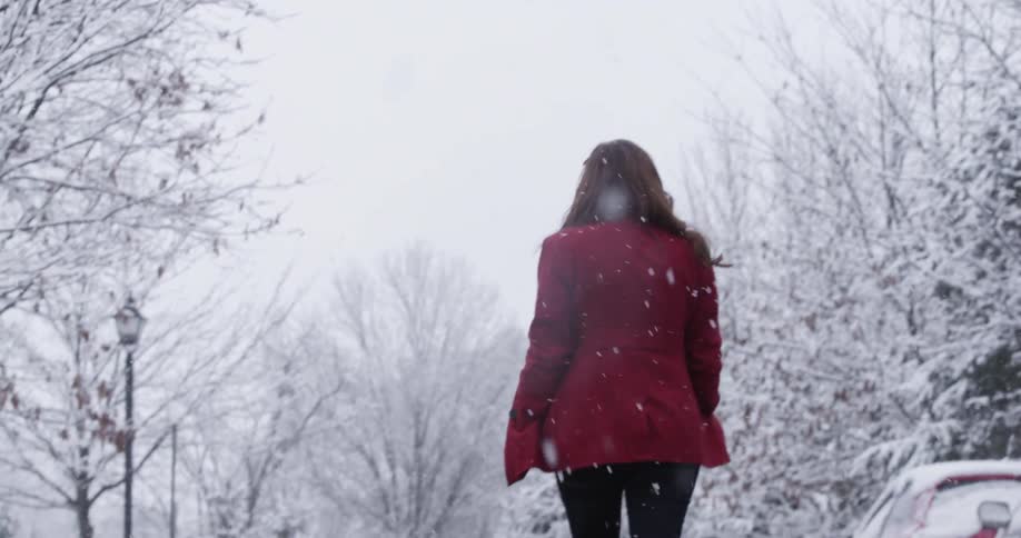 A young woman wearing a coat twirls around happily outdoors while snow is falling
