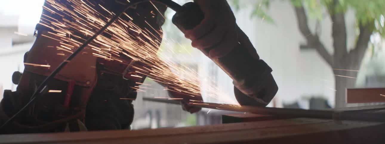A man uses a small handheld buzz saw to cut through a piece of a steel pipe outdoors at daytime