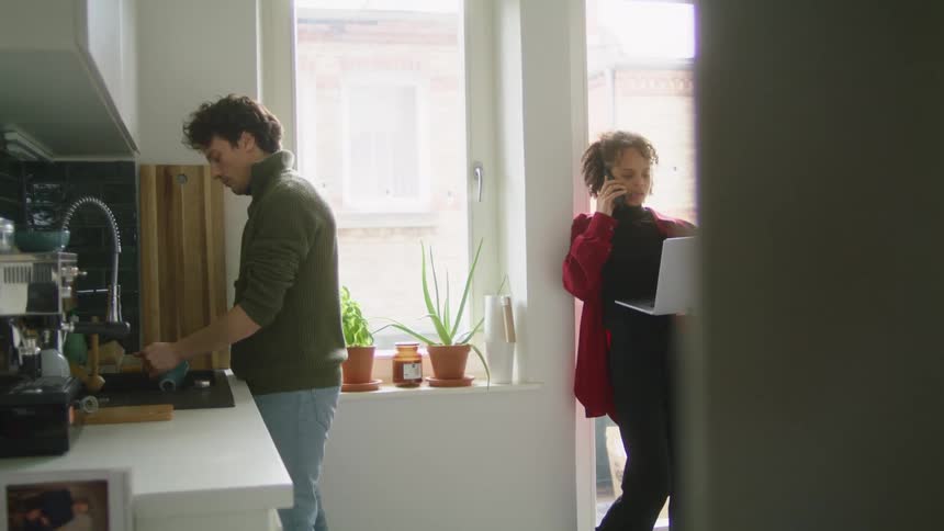 A couple in a kitchen with the man washing dishes in the sink and the woman standing up against a window holding a laptop while talking on the phone at daytime
