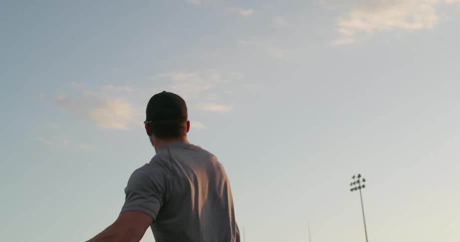 A man with a baseball hat and baseball glove throwing a baseball on a baseball field with the sky and clouds in the background