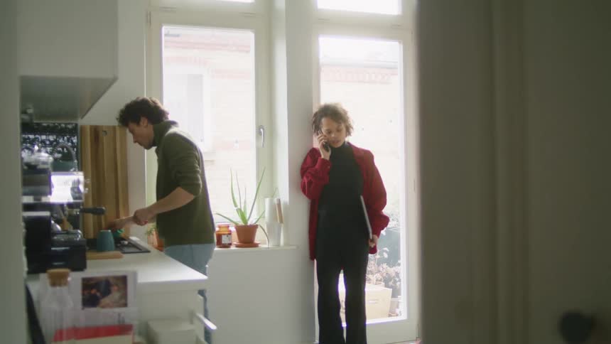 A woman on a phone holding a laptop leans against a door with a window where the sun is shining through in a kitchen next to a man cleaning a stove top