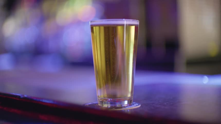 A full glass of beer sits on a coaster at the edge of a bar