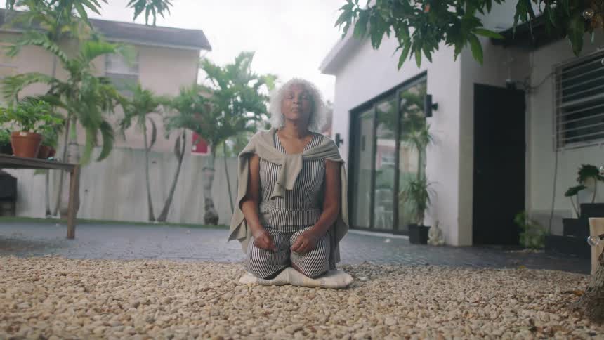 A woman kneels on the ground on her patio with her eyes closed while meditating