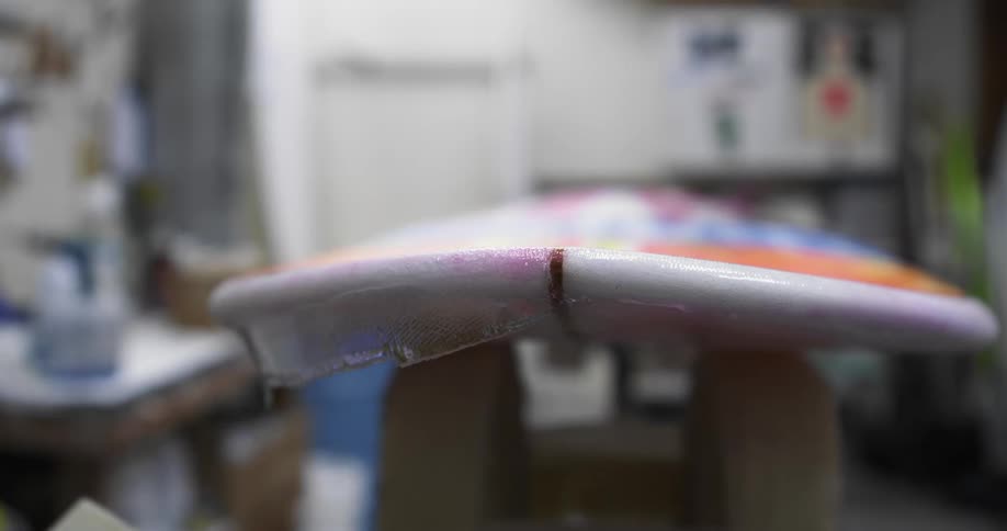 A person applies a layer of clear film to a surfboard with a spatula in a workshop