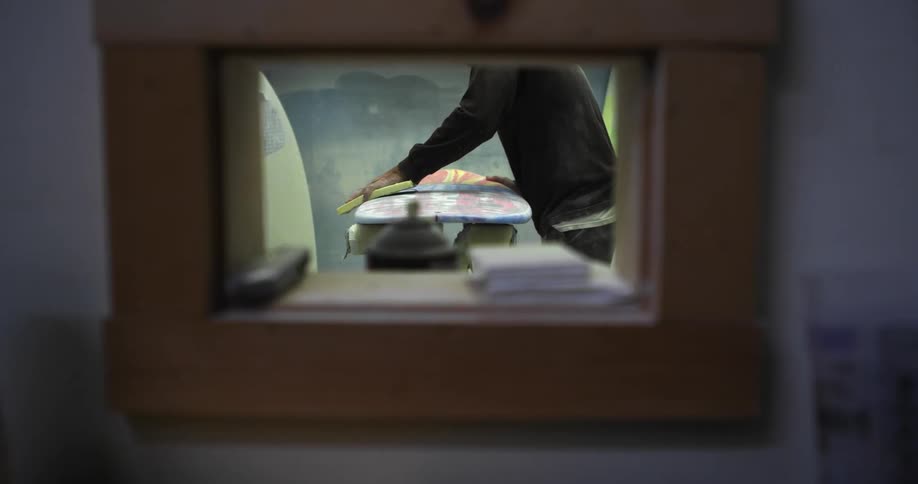 A small window shows a man cleaning a surfboard in another room