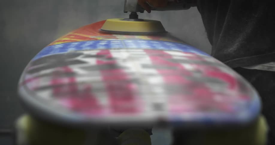 A person uses a power tool to sand the surface of a surfboard inside a workshop