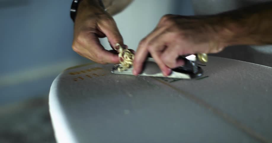 A man uses a hand planer to smooth the surface of a surfboard he is repairing