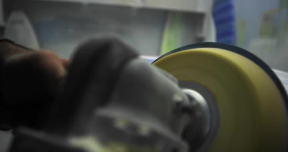 Man using a yellow spinning polishing tool to smooth the edges of a surfboard on a workshop table