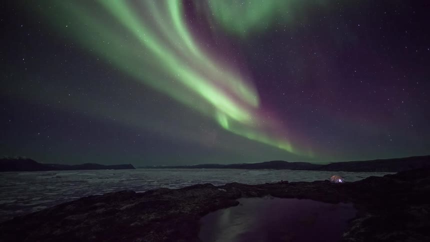 The northern lights shimmer and wave in the sky above icy water and a glowing tent campsite