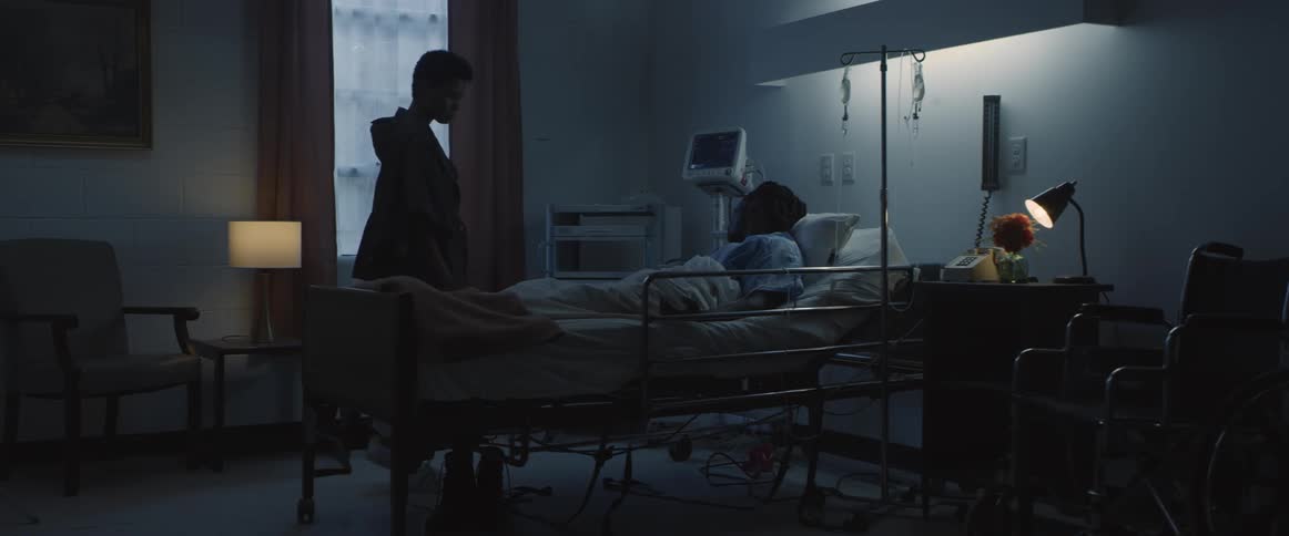 A woman walking up and kneeling down next to a person on a hospital bed with a wall and window in the background