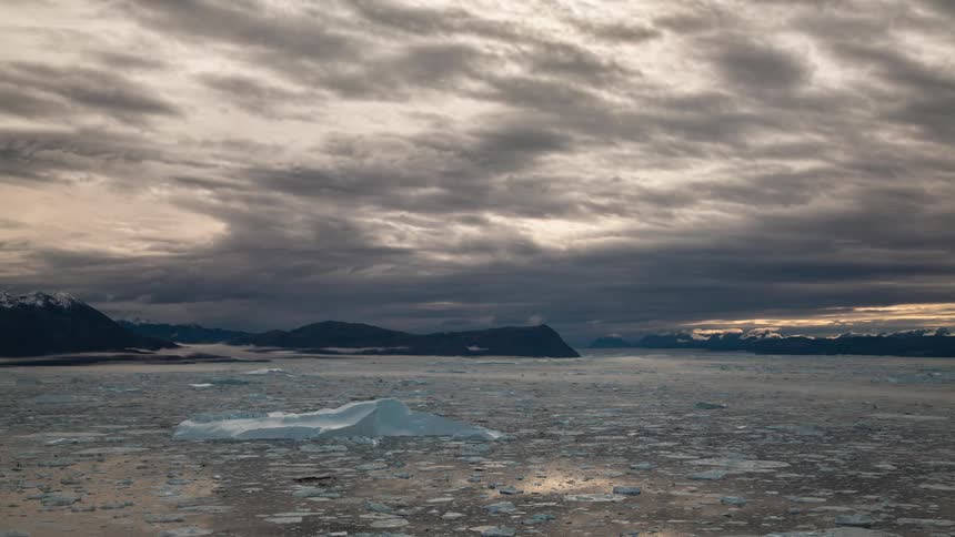 A lake with water and ice with a mountain range the sky and clouds in the background