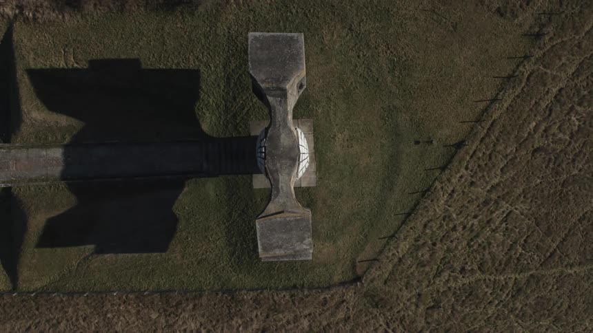 A large hammer like monument casts a shadow onto the grass in a rural field