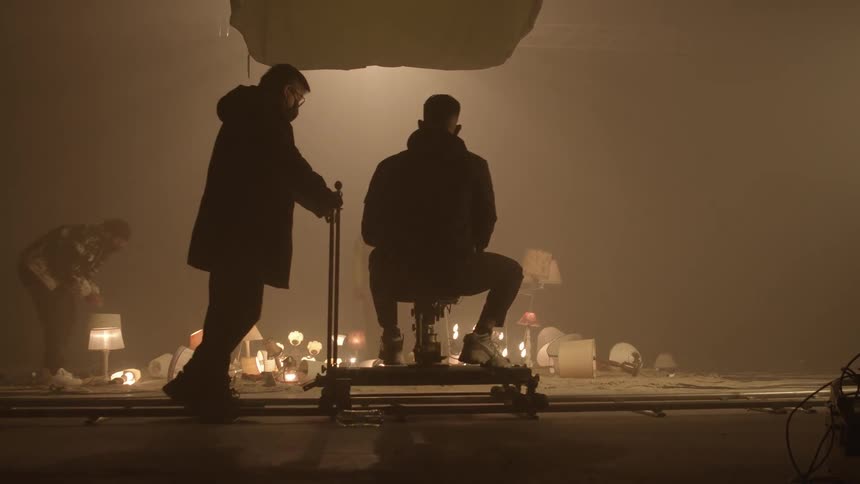 A man pushes a film maker on a cart as he films people on a stage looking at lamps