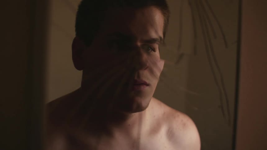 A man in a room standing and looking at his reflection on a broken mirror