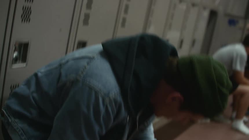 A teenage boy walks over to a bench in front of lockers and places a bag and some equipment down and then sits down while two other boys talk