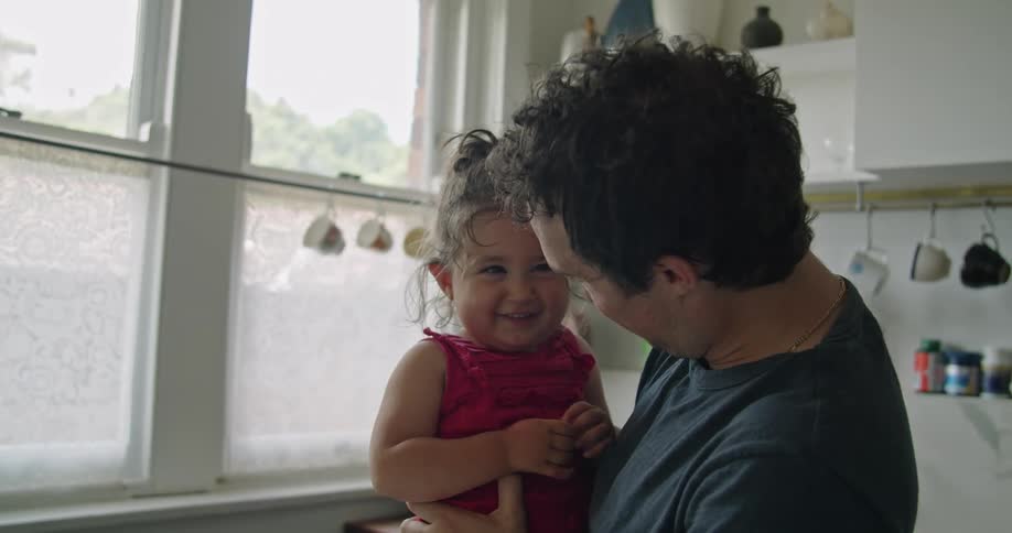 A father standing in a kitchen at home at daytime holding his daughter looks her in the face and she smiles