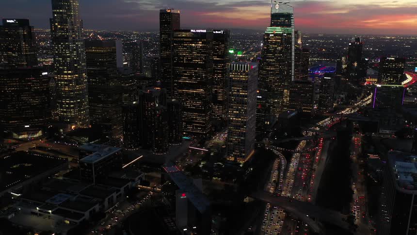 Heavy traffic passes through a downtown city filled with tall skyscrapers lit up as the sun sets
