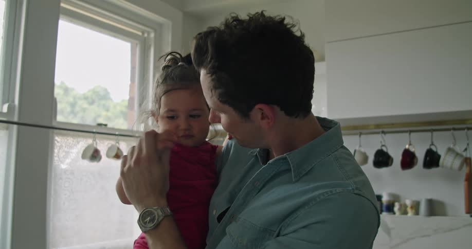 A man in a kitchen holding a baby girl while looking at a laptop on a table