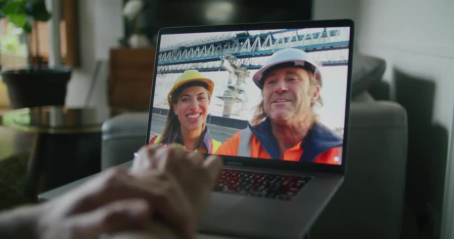 Two construction workers wearing hardhats on a video call in a laptop with someone at home