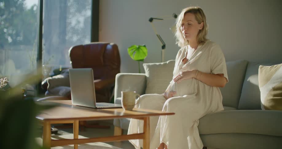 Blonde pregnant woman with white dress makes a video call on laptop in the living room at home