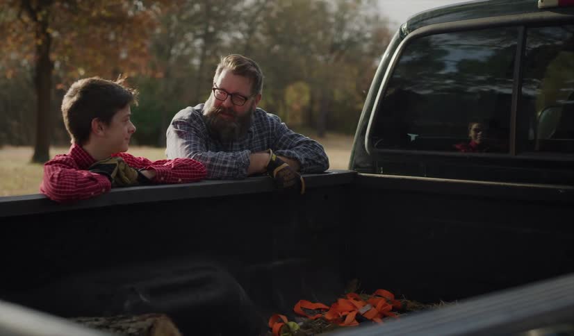 A dad pats his son on the back as they lean against the pickup truck and talk