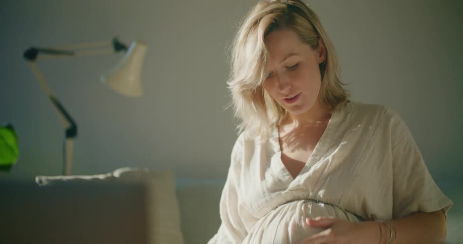 Blonde pregnant woman with white dress and bracelet makes video call on laptop in the living room