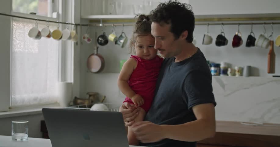 A man in a kitchen holding a baby girl while looking at a laptop on a table