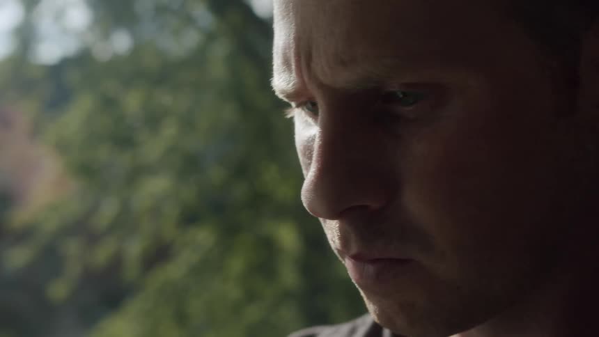 A man turns his head from looking out of a window and stares off in front of him with a gloomy expression