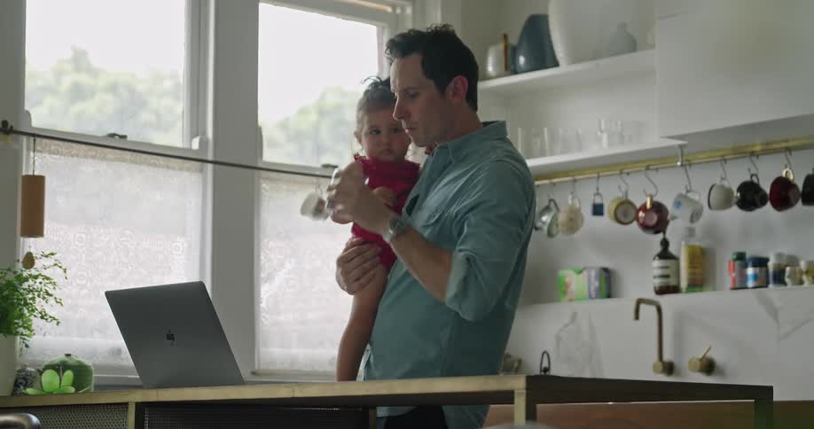 Father holding daughter in his arms drinks water while using a laptop in the kitchen