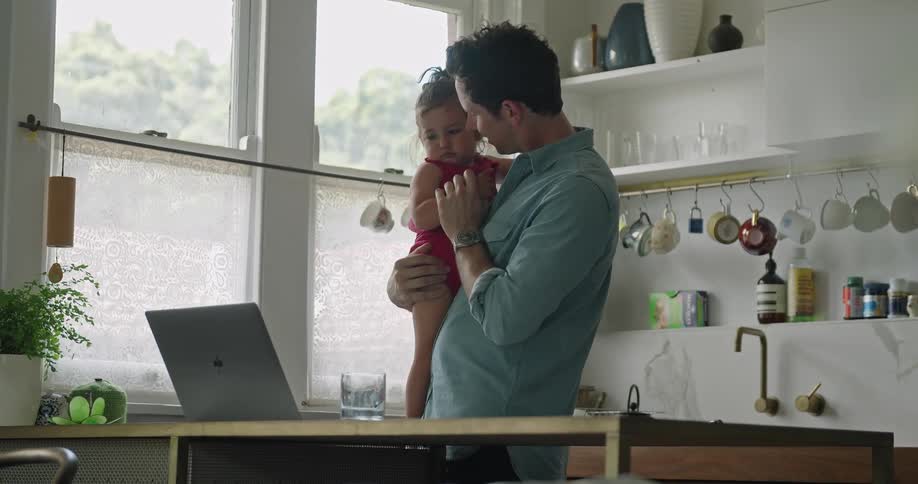 Father standing in a kitchen during the day holding his daughter in his arms and bouncing her while waving at a laptop camera