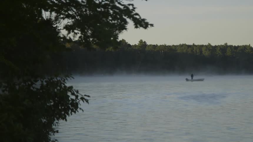 A person standing in a small boat fishes as fog rises from a lake at dawn