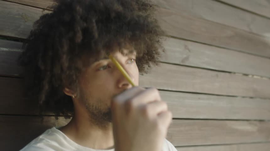A man sitting up against the side of a wooden building with a pondering expression and taps a pencil against his head then starts writing