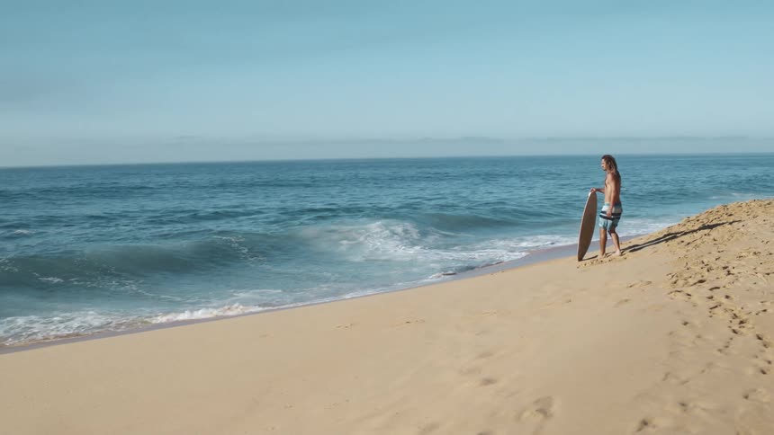 A man is standing on a beach holding his surfboard and looking at the waves