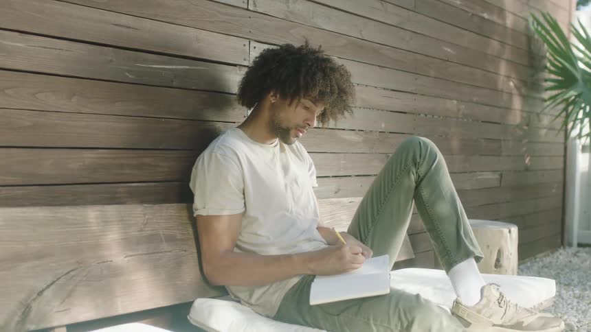A man sitting next to a building writing on a book with a pencil