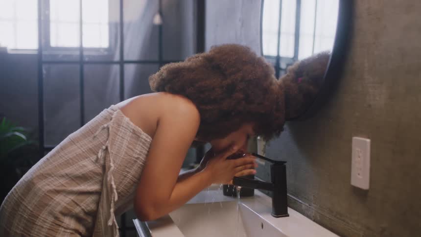 Brunette woman with frizzy hair washes her face and looks in the mirror in the bathroom