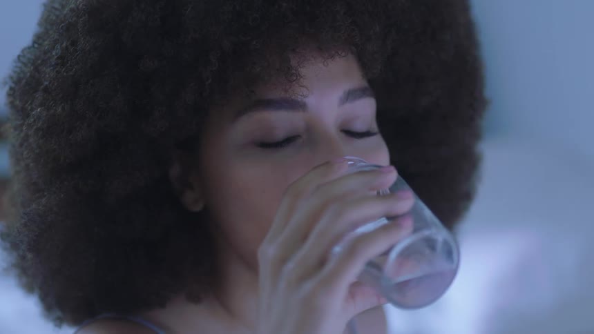 Young woman with an afro drinking water from a small glass bottle at home