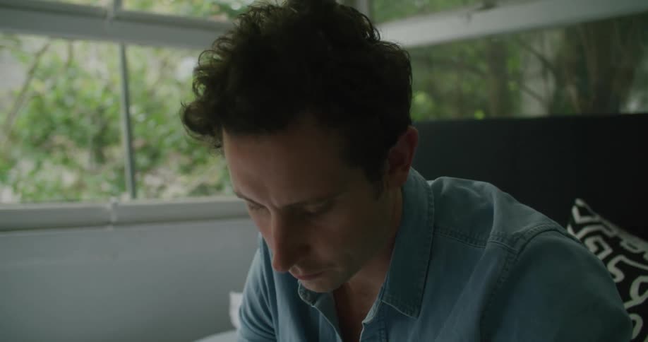 Man with blue shirt sits on bed and works on laptop in the bedroom